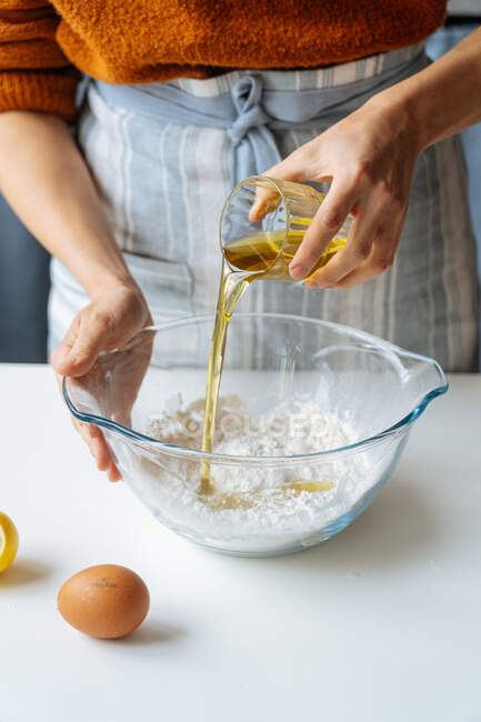 Сельскохозяйственная женщина наливает оливковое масло из стекла в большую миску с мукой во время приготовления теста на белом столе на кухне — стоковое фото