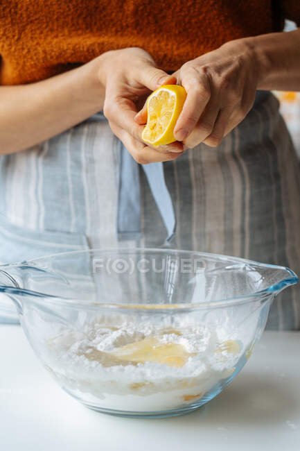 Crop casual female in apron squeezing lemon over glass transparent bowl with mixed ingredients while preparing dough at home kitchen — Stock Photo