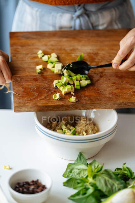 Ritagliare femmina tenendo tagliere di legno e l'aggiunta di avocado tritato alla ciotola di ceramica con quinoa durante la preparazione di cibo in cucina a casa — Foto stock