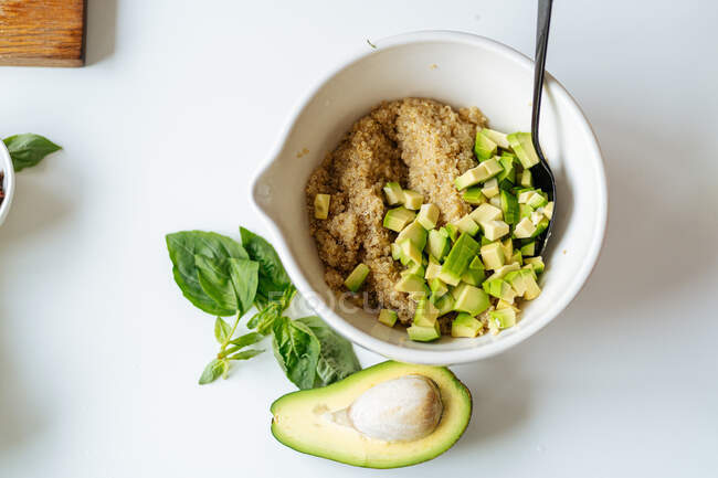 Dall'alto di ciotola in ceramica bianca con quinoa e avocado tritato posto su tavolo bianco con metà di avocado e foglie di basilico verde — Foto stock