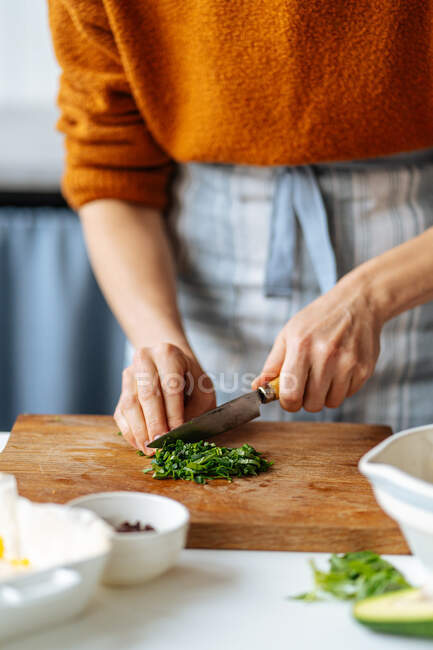 Crop housewife cutting fresh green herbs on wooden board while preparing delicious dinner at home kitchen — Stock Photo