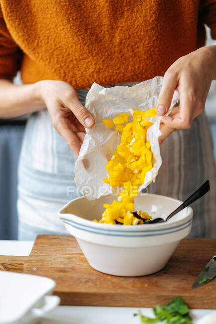 Crop casalinga mettendo mango tritato in ciotola con ingredienti alimentari collocati sul tagliere di legno durante la preparazione della cena a casa — Foto stock