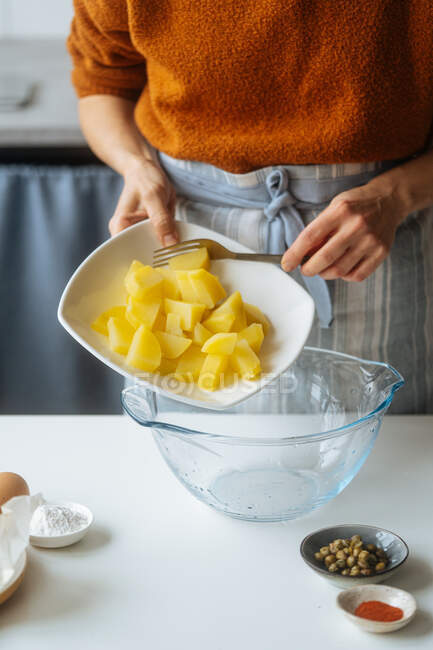 Cultiver la femelle en mettant les pommes de terre cuites coupées dans un bol en verre tout en préparant la nourriture à la table blanche à la cuisine — Photo de stock