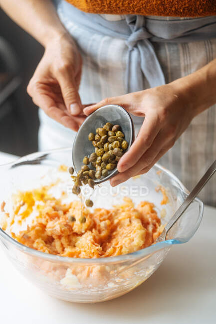 Cosecha hembra añadiendo alcaparras marinadas en un tazón de vidrio transparente con relleno de salmón mientras preparas una cena saludable en la cocina casera - foto de stock