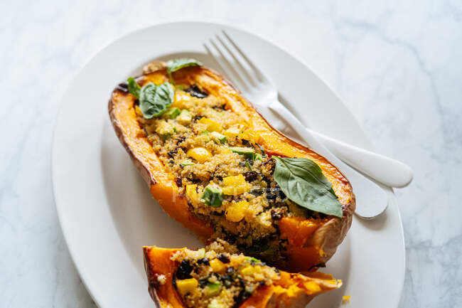 Closeup of delicious appetizing vegetarian stuffed butternut squash with quinoa and vegetables served on white plate with fork and knife on marble background — Stock Photo