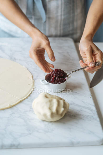 Von oben geerntet fleißige Köchin nimmt appetitliche Marmelade mit Löffel in Teig für die Zubereitung von süßem Gebäck auf Marmortisch in heller Küche gelegt — Stockfoto
