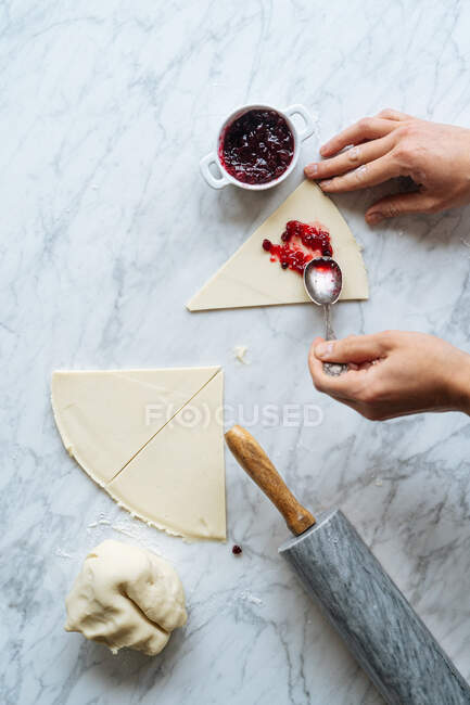 De arriba cocine la cosecha colocando diligentemente mermelada con cuchara sosteniendo la masa a mano en la mesa con masa y rodillo en la cocina - foto de stock