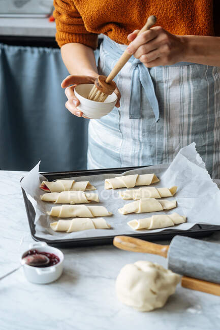 Cozinhe tigela segurando e escovando croissants na assadeira na mesa — Fotografia de Stock