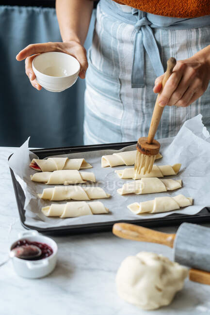 Von oben hält Koch weiße Schüssel in der Hand und bürstet fleißig leckere Croissants auf Backblech auf dem Tisch in der Küche — Stockfoto