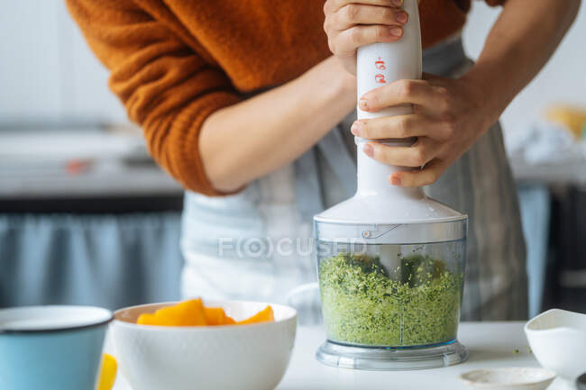 Crop cook preparing food and mixing green vegetables in white blender on table in light kitchen — Stock Photo
