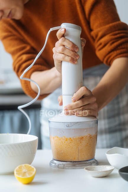 Crop cook mixing pumpkin and checking bowl holding blender in hand on table with citrus in light kitchen — Stock Photo