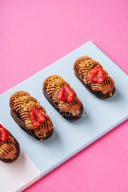 From above delectable chocolate eclairs with pieces of fresh raspberries and caramel cream placed in line on board on pink background — Stock Photo