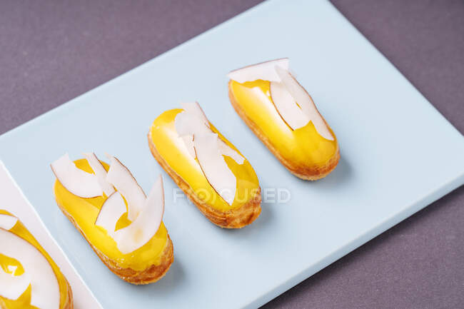 Primeros planos sabrosas eclairs con glaseado de plátano y trozos de coco colocados sobre tabla azul - foto de stock