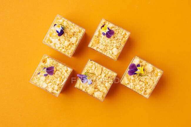 Vue de dessus des tasses carrées avec de délicieuses pâtisseries décorées de fleurs et placées sur fond orange — Photo de stock