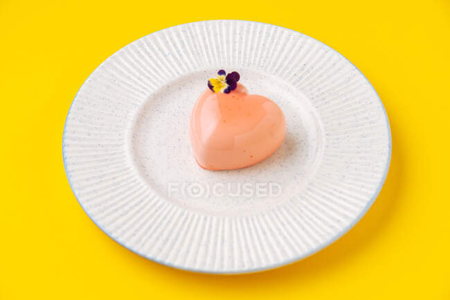 From above small heart shaped dessert with pink icing and small flower placed on plate on yellow background — Stock Photo