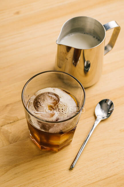 From above of stylish glass with ice black coffee on wooden table with milk pitcher and spoon — Stock Photo