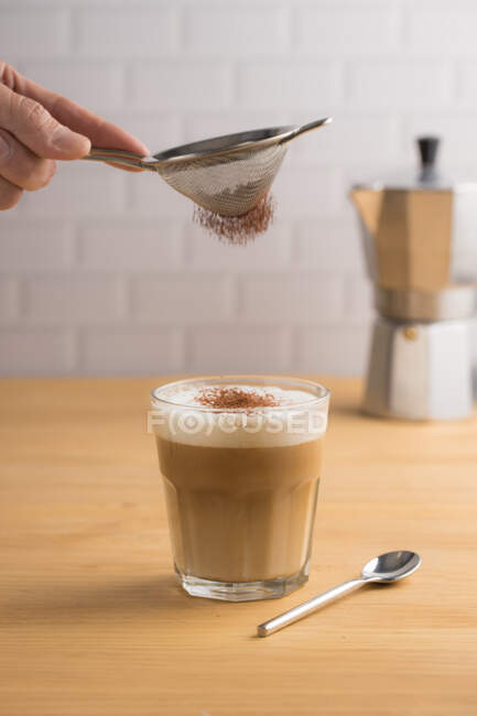 Anonymous barista sifting cacao powder by sieve on glass with brewed coffee and milk foam in cafe — Stock Photo