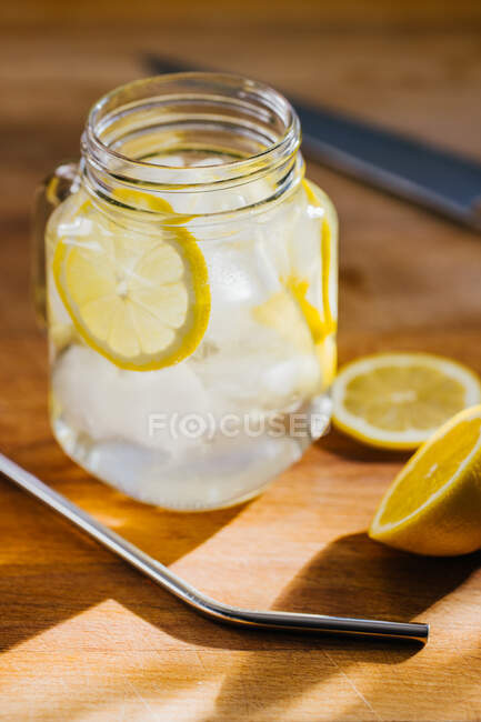 De arriba metálico reutilizable paja y jarra de vidrio con hielo y rodajas de limón en la mesa de madera en la cocina - foto de stock
