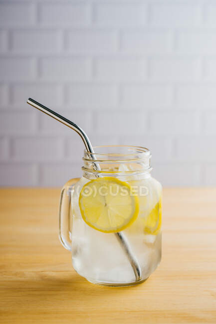 Jarra metálica reutilizable de paja y vidrio con rodajas de hielo y limón sobre mesa de madera en cocina - foto de stock
