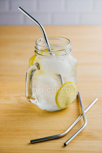 Metallwiederverwendbare Stroh- und Glaskanne mit Eis- und Zitronenscheiben auf Holztisch in der Küche — Stockfoto