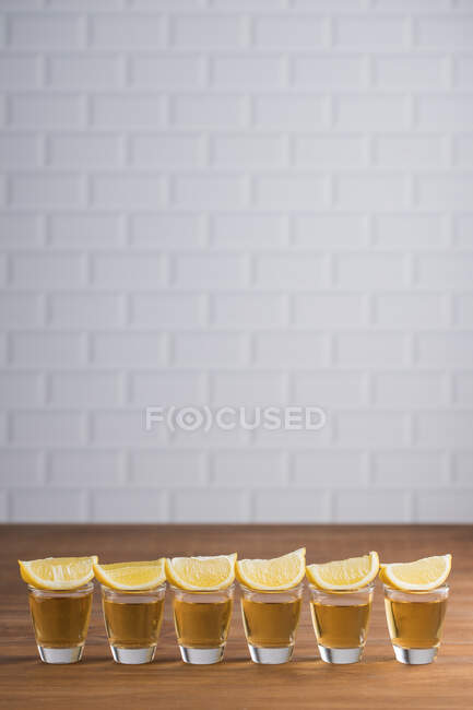 Reihe von Glasschlitzen mit goldenem Tequila und Zitronenscheiben auf Holztisch mit weißer Wand auf verschwommenem Hintergrund — Stockfoto