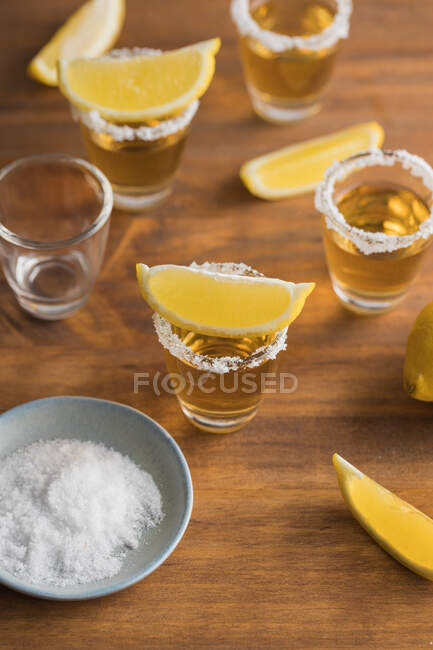 De cima vista superior de tiros de vidro de tequila dourada com borda salgada e fatias de limão em cima na mesa de madeira — Fotografia de Stock