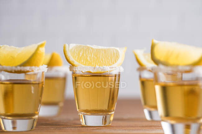 Fila de cerdas de vidrio con tequila dorado y rodajas de limón sobre mesa de madera con pared blanca sobre fondo borroso - foto de stock