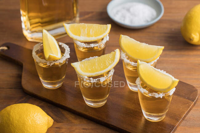 De acima mencionados tiros de vidro de tequila dourada com borda salgada e fatias de limão em cima na mesa de madeira — Fotografia de Stock