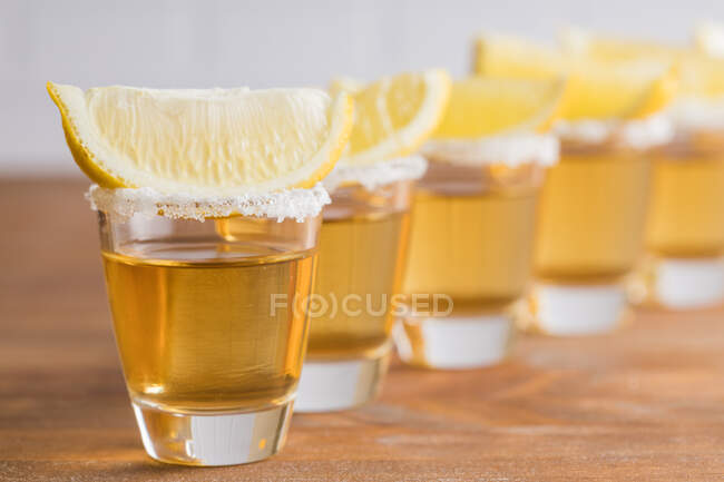 Fila de sabões de vidro com tequila dourada e fatias de limão na mesa de madeira com parede branca no fundo embaçado — Fotografia de Stock