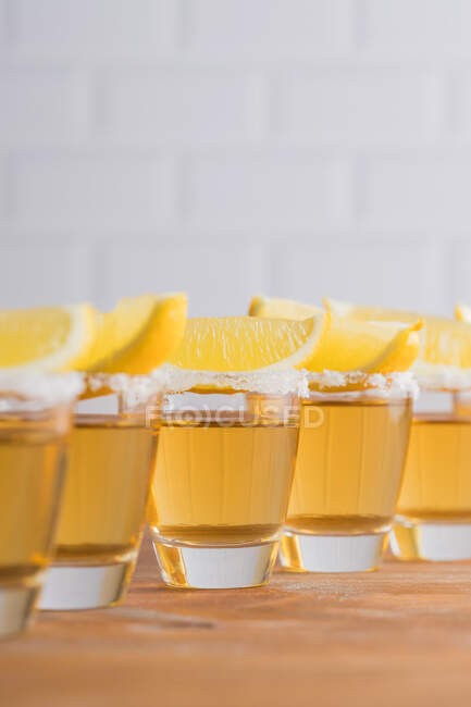 Row of glass sots with golden tequila and slices of lemon on wooden table with white wall on blurred background — Stock Photo