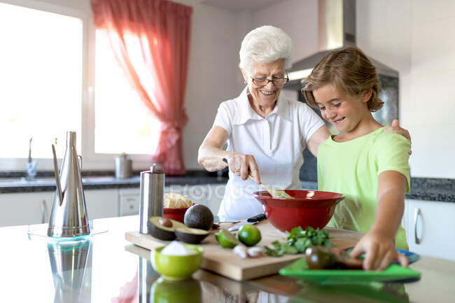 Fröhliche alte Frau mit weißem Haar hilft Kind beim gemeinsamen Zubereiten von Guacamole in der Küche — Stockfoto