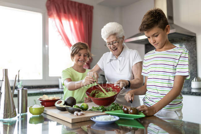 Cheerful old woman with white hair helping children while preparing guacamole together in kitchen — Stock Photo