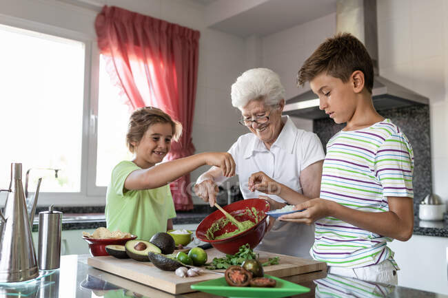 Fröhliche alte Frau mit weißem Haar hilft Kindern beim gemeinsamen Zubereiten von Guacamole in der Küche — Stockfoto
