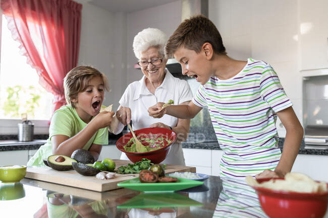 Cheerful grandma and boys tasting homemade guacamole paste with tortilla chips in kitchen — Stock Photo
