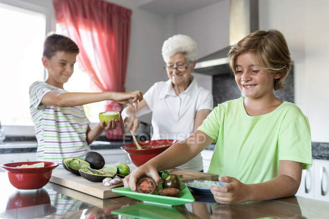 Mulher idosa alegre com cabelos brancos ajudando crianças enquanto prepara guacamole juntos na cozinha — Fotografia de Stock