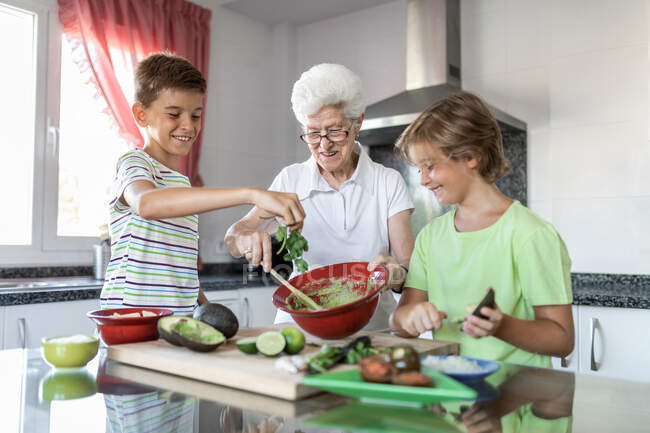 Allegro vecchia donna con i capelli bianchi aiutare i bambini mentre si prepara guacamole insieme in cucina — Foto stock