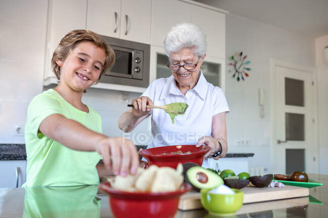 Веселая старая женщина с седыми волосами помогает ребенку во время приготовления гуакамоле вместе на кухне — стоковое фото