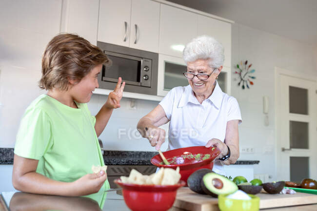 Весела стара жінка з білим волоссям допомагає дитині під час підготовки гуакамоле разом на кухні — стокове фото