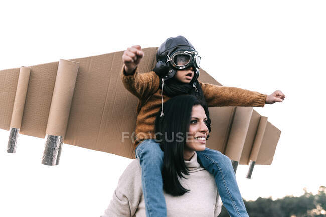 Glücklicher süßer Junge mit Brille und Pappflügeln, während er auf den Schultern seiner Mutter sitzt und Flieger auf einer Wiese im Gegenlicht imitiert — Stockfoto