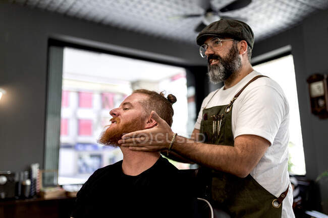 Vista laterale del barbiere che fa massaggio al viso al bel rossa uomo con seduta in sedia — Foto stock