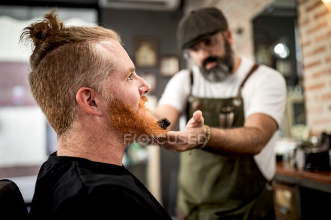 Peluquero con recortadora cortando la barba del pelirrojo sentado en la peluquería - foto de stock