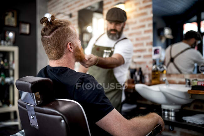 Barbeiro aparando barba de homem ruivo sentado na barbearia — Fotografia de Stock