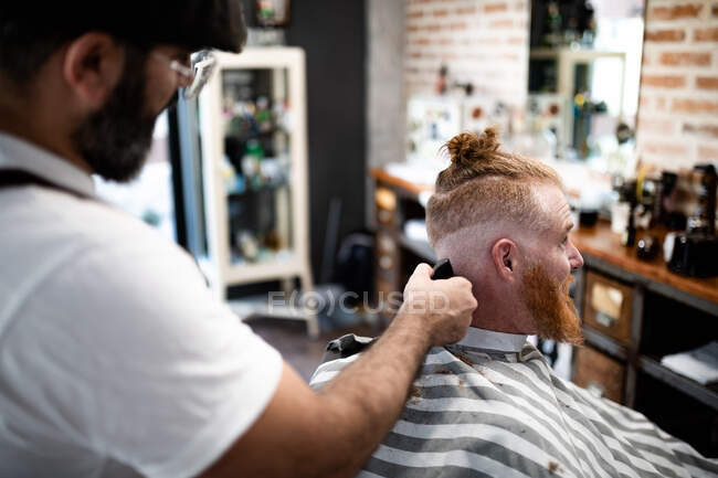 Современный парикмахер, стригущий волосы взрослого мужчины в кресле парикмахера — стоковое фото