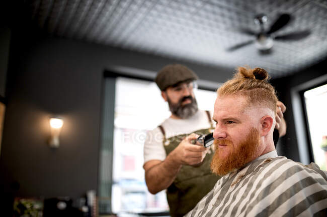 Modern hairdresser barber cutting a adult man redhead's hair in barber chair — Stock Photo