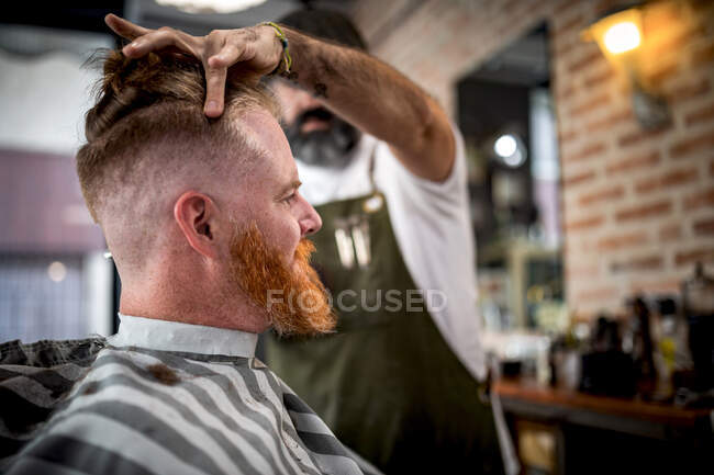 Vue de côté coiffeur moderne coupant les cheveux d'un homme adulte rousse dans la chaise de coiffeur — Photo de stock