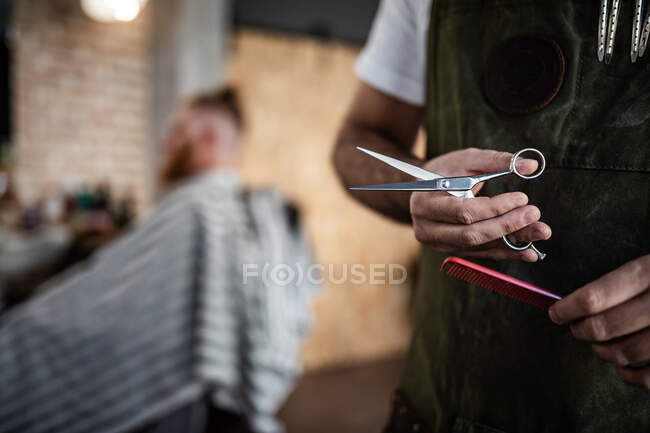 Unrecognizable crop man hairdresser holding scissors and comb in modern barbershop — Stock Photo
