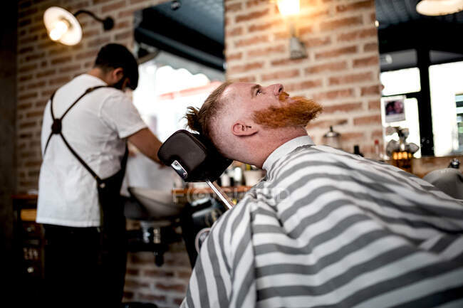 Pelirrojo sentado en la peluquería esperando al barbero anónimo en el fondo - foto de stock