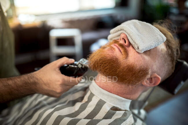 Peluquero irreconocible recortado con recortadora barba pelirroja con toalla cubriendo los ojos sentado en la barbería - foto de stock