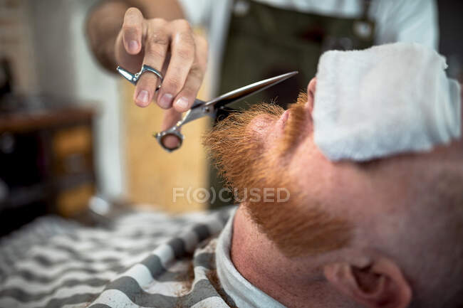 Обрезанный неузнаваемый парикмахер с ножницами, срезающий бороду рыжеволосого человека, сидящего в парикмахерской с глазами, покрытыми полотенцем — стоковое фото