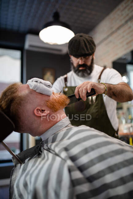 Peluquero borroso irreconocible con peine que fija la barba pelirroja con toalla que cubre los ojos sentado en la barbería - foto de stock